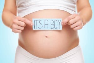 A pregnant person holds a blue sign reading IT'S A BOY in front of their bare belly. The person is wearing a white top pulled up to expose the belly and white bottoms, showcasing one of the Signs You’re Having a Boy. The background is light blue.