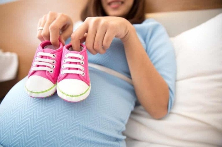 A woman wearing a light blue dress holds a pair of small, pink baby shoes above her pregnant belly, hinting she might conceive a girl. She is sitting on a white bed, with part of her face visible in the background.