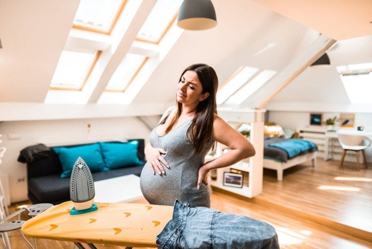 A pregnant woman stands in a room with her hand on her back, looking tired, possibly experiencing round ligament pain. An ironing board with a pair of jeans and an iron is in front of her. The room features skylight windows, a bed, a couch, and a desk.