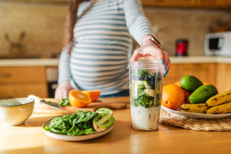 A person in a striped shirt is preparing a smoothie in a kitchen. The blender container contains various ingredients, including greens and fruits like spinach, kiwi, avocado, bananas, and oranges—perfect for those curious about how to prevent gestational diabetes through a balanced diet.