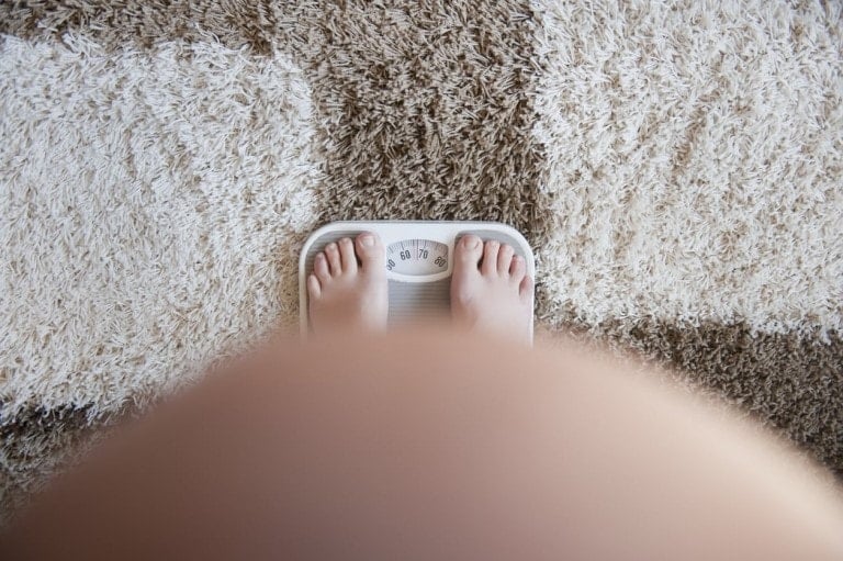 A person is standing barefoot on a scale, which shows a weight measurement of around 86 kilograms. The viewpoint focuses on the scale and feet, with part of the person's body visible in the foreground. This could be part of tracking pregnancy weight gain, as the scale rests on a textured, multi-colored rug.
