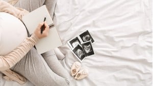 A pregnant person is sitting on a bed holding a pen and a notebook, jotting down ideas as they try to agree on a baby name. Ultrasound images and a pair of baby shoes are beside them on the bed. Their hands are resting on their belly, and the bed is covered in white sheets.