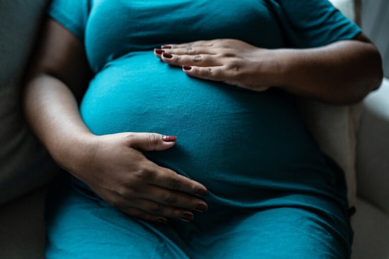 A close-up of a pregnant person, experiencing a geriatric pregnancy, sitting on a couch in a teal dress. Their hands, adorned with painted nails, rest gently on their large stomach. The scene is indoors with soft lighting.