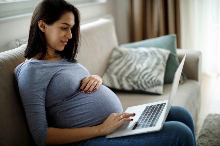A pregnant woman is sitting on a couch, holding her belly with one hand and using a laptop with the other, possibly writing a Babylist baby registry review. She is dressed in a gray long-sleeve shirt and blue jeans. The background includes pillows and a window with curtains.