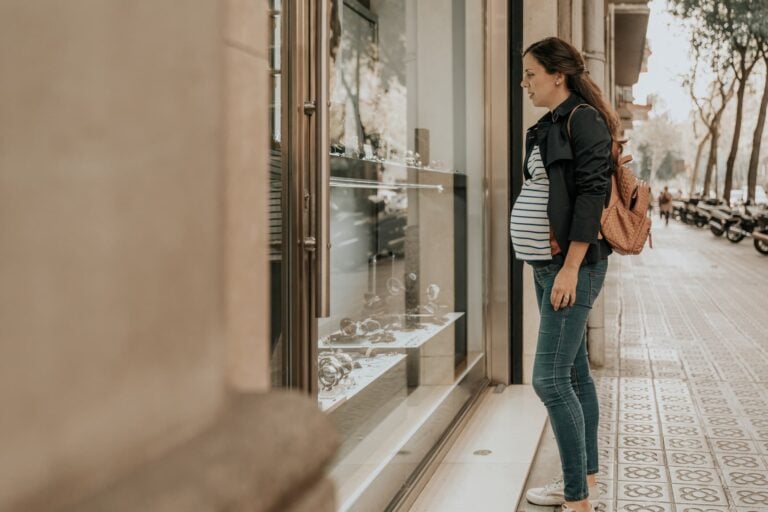 A pregnant woman stands on a city sidewalk, looking at items displayed in a shop window. She is wearing a black jacket, striped shirt, blue jeans, and comfortable shoes during pregnancy—white sneakers—with a brown backpack on her shoulder. Trees and parked motorcycles line the street.