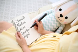 A person in a yellow outfit is sitting and holding a notebook in their lap. They are writing down Southern girl names such as Charlotte, Madison, Alison, Scarlett, and Abigail with a black marker. Next to them is a stuffed animal dog wearing a zigzag-patterned shirt.