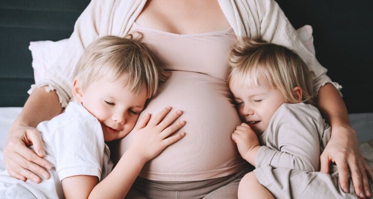 A pregnant woman sits with her two young children, enjoying the closeness that thoughtful pregnancy spacing can bring. Both kids rest their heads on her belly, one on each side, smiling with closed eyes. The woman is wearing a light pink top and a white cardigan.