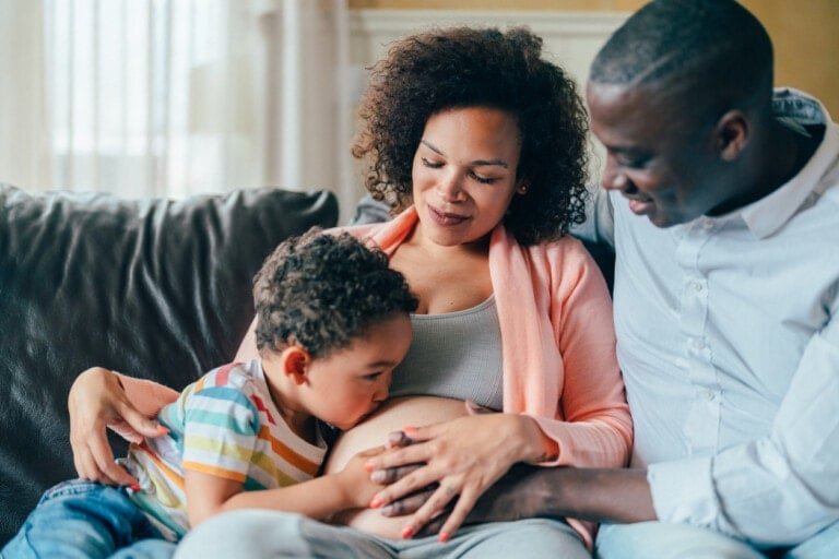 A pregnant woman sits on a couch next to a man while a young boy kisses her belly and rests his hands there. The man places a hand on the boy's back, looking at the woman's belly with a smile. This family is joyfully expecting their second child.