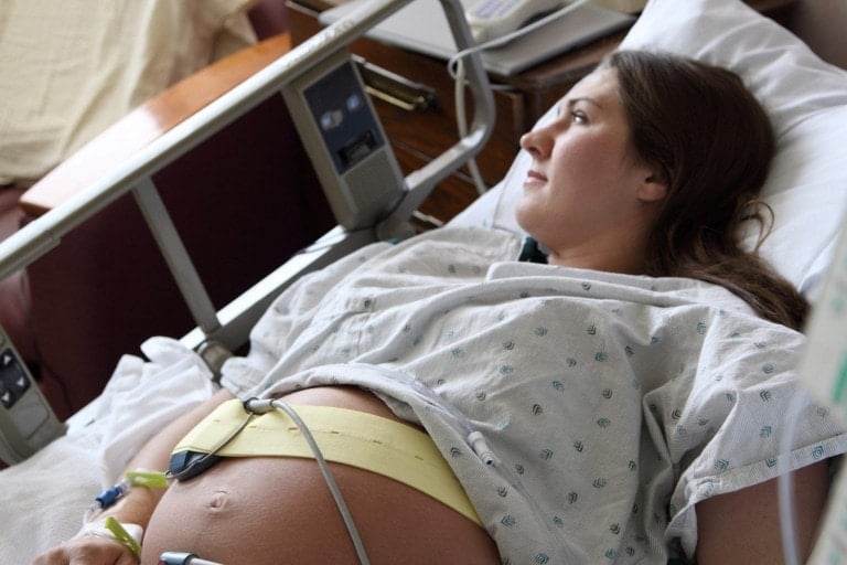 A pregnant woman lying in a hospital bed, hooked up to monitoring equipment, appears to be thoughtfully looking to her left. She wears a hospital gown with sensors attached to her belly, likely for fetal monitoring. Perhaps she's reflecting on a recent episode of the Go to the Hospital in Labor podcast.