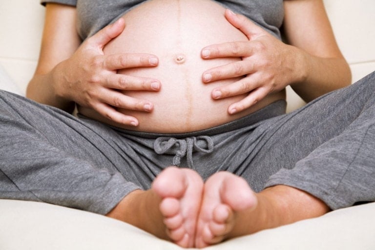 A pregnant person wearing grey clothing sits cross-legged, holding their bare, visibly pregnant belly with both hands. The focus is on the abdomen and the prominent linea nigra running down the center of their stomach. Their feet are pointing towards the camera.