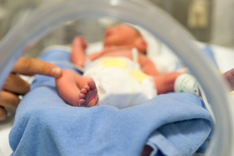 A newborn baby in the NICU lies on a blue blanket inside an incubator. An adult's finger gently touches the baby's foot. The baby is wearing a diaper and has a hospital wristband. The background is out of focus, emphasizing the baby and the interaction.