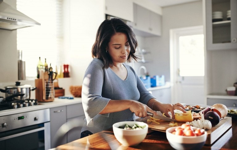 A woman is slicing vegetables on a wooden cutting board in a modern kitchen. Various chopped vegetables and utensils are on the counter as she focuses on her meal planning, with natural light coming in through the windows.