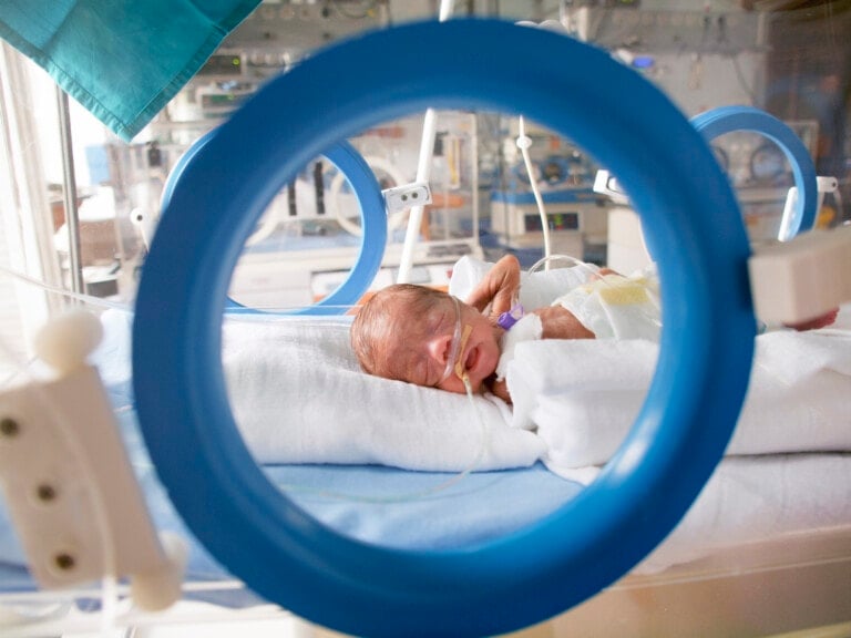 A premature baby lies in an incubator in a neonatal intensive care unit. The baby is connected to medical tubes and monitors, and is wrapped in white blankets. The image, possibly reflecting concerns about the U.S. Premature Birth Rate, is seen through a circular opening, likely part of the equipment.