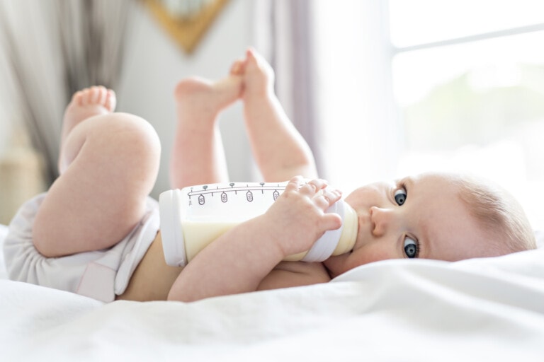 A baby lies on a bed, wearing a diaper and holding a baby bottle with both hands. The baby's feet are raised in the air as they enjoy feeding from the bottle. The background features a softly lit window and blurred decor.