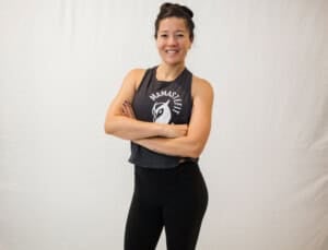 A person stands against a plain background with arms crossed, smiling. They wear a sleeveless black shirt with a graphic design and black leggings.