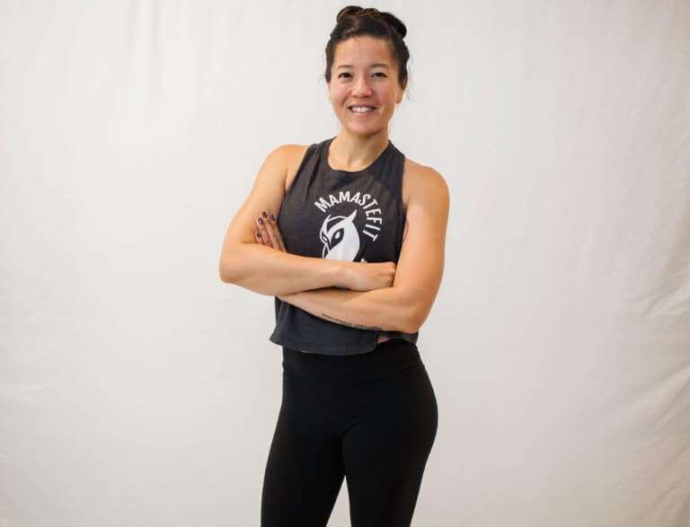 A person stands against a plain background with arms crossed, smiling. They wear a sleeveless black shirt with a graphic design and black leggings.