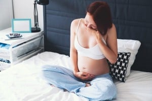 A pregnant person sits on a bed, dressed in a white sports bra and blue striped pajama pants. They are touching their belly with one hand and covering their mouth with the other, displaying early pregnancy signs. A bedside table in the background holds some items.