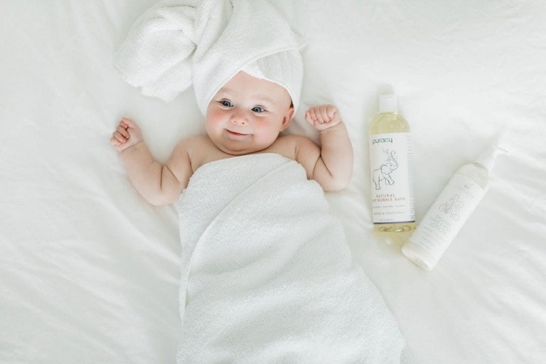 A baby is lying on a white surface, wrapped in a white towel with a smaller towel around their head. Two bottles of Puracy baby skincare products, known for their natural cleaning properties, are placed beside the baby. The little one has a content expression and is looking at the camera.