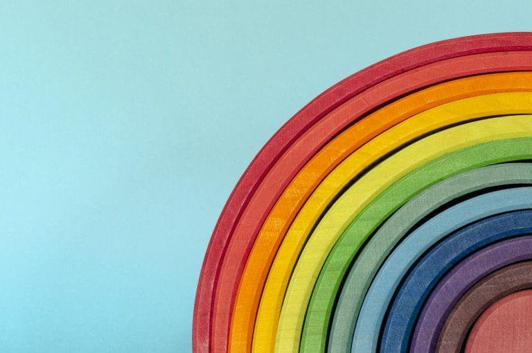 A close-up of a wooden rainbow stacker toy against a light blue background. This Montessori toy showcases various colors in concentric arch shapes, including red, orange, yellow, green, light blue, dark blue and purple, arranged from the outermost to the innermost layers.