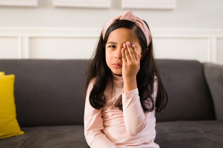 A young girl with long dark hair, wearing a pink headband and a pink long-sleeve shirt, covers one eye with her hand while sitting on a gray sofa. A yellow cushion rests beside her. Her posture suggests she might be experiencing discomfort, possibly from pink eye in kids.