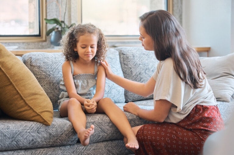 A woman is sitting on a couch next to a young girl with curly hair. The girl looks upset and has her hands clasped in her lap. The woman has her hand gently placed on the girl's shoulder, possibly offering comfort and compassionately discipline. They are indoors with a window in the background.