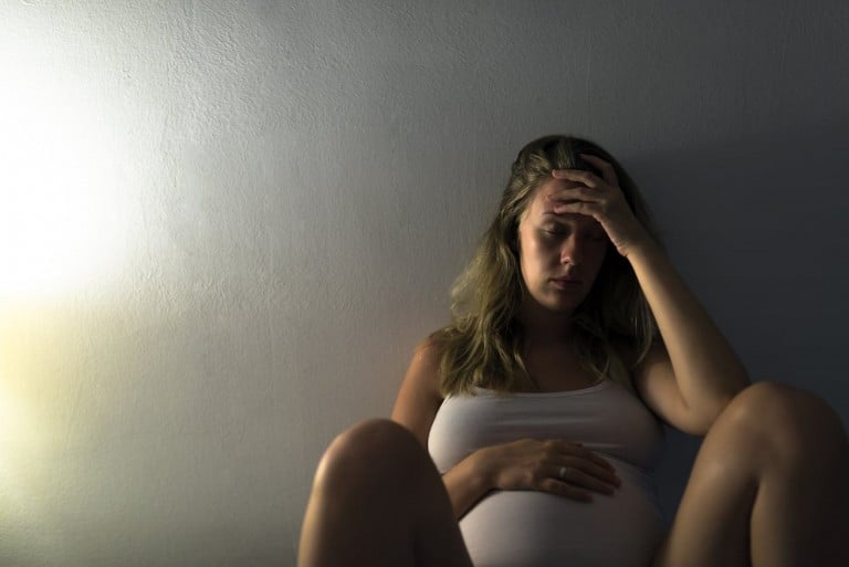 A pregnant woman in a white tank top sits against a wall with her legs bent. She places her left hand on her stomach and her right hand on her forehead, appearing distressed from financial stress while pregnant. The lighting is dim, with a lamp on the left side casting a soft glow.