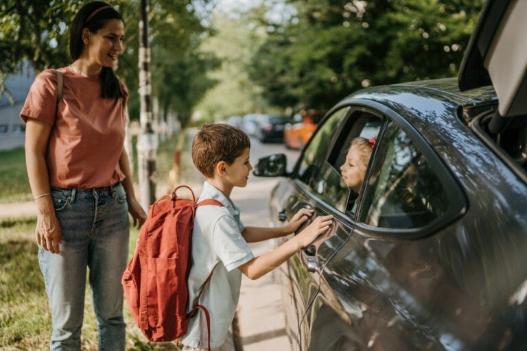 A woman and a young boy with a red backpack stand near a car. The boy touches the car window, interacting with his kid's friend's parents inside. The car is parked on a tree-lined street. The woman is smiling and looking at the children.