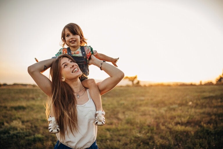 A woman with long hair, wearing a white sleeveless top, is giving a piggyback ride to a young child. The child is smiling, wearing a colorful shirt and overalls. They are outdoors in a grassy field at sunset, enjoying some mental health activities for kids amidst the golden glow.
