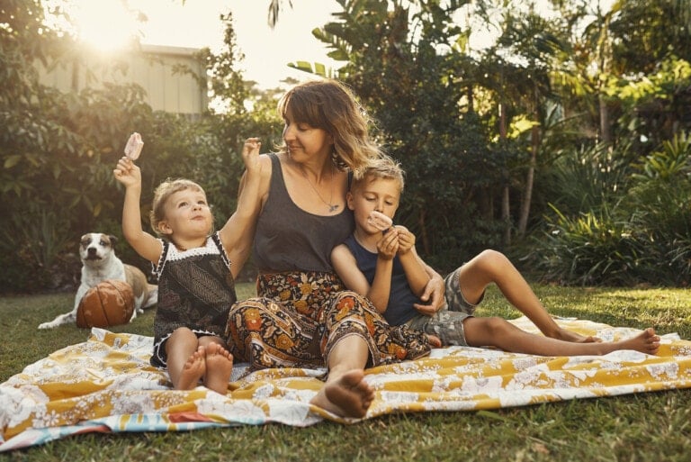 A woman sits on a picnic blanket in a garden with two children, whom she loves dearly. One child is holding a popsicle, and the other is eating a cookie. A dog sits beside them with a ball. Trees and shrubbery can be seen in the background. The sun is shining brightly.