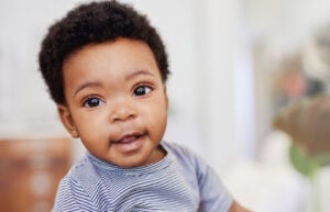 A baby with short curly hair is looking at the camera with a neutral expression, embodying one of those 11-month milestones. The baby is wearing a striped shirt. The background is blurred, which emphasizes the baby's face.
