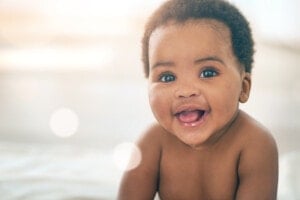 A smiling baby is shown in close-up, looking directly at the camera with an open mouth. The baby, likely to be adorned with traditional African girl names, has curly hair and bright eyes. The background is softly blurred with warm and gentle light, capturing the baby’s cheerful and happy mood.