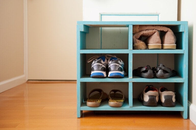 A small, turquoise shoe rack stands empty against a white wall on a hardwood floor. Though there are no shoes inside, its design hints at an organized display for athletic sneakers, sandals, formal shoes, and boots.