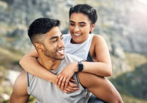 A couple outdoors, with a woman playfully getting a piggyback ride from a man. They are both smiling in an open, mountainous setting. The woman is wearing a smartwatch.