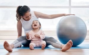 Here is your text with all single and double quote characters removed: A woman sits on a yoga mat with a baby in her lap, both barefoot. The woman is smiling and touching a large exercise ball beside them. The baby is wearing a pink outfit and looking up with an open mouth.