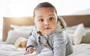 A baby with a focused expression is crawling on a bed. The boy, possibly destined for one of those strong T names, is wearing a grey, hooded sweater. The background includes white bedding and a headboard, with a soft light coming from the left side.