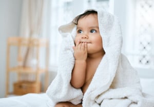 A baby is wrapped in a white towel with a hood, sitting on a bed. The baby has one hand near its mouth and gazes upward, like they're pondering old Persian names. Daylight streams through the windows in the background.