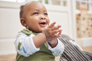 A 7-month-old baby wearing a long-sleeved shirt with colorful patterns and a green vest is sitting and clapping hands. The baby is smiling and looking up. The background is indoors with light walls and windows, creating a cheerful atmosphere during the baby's wake window.
