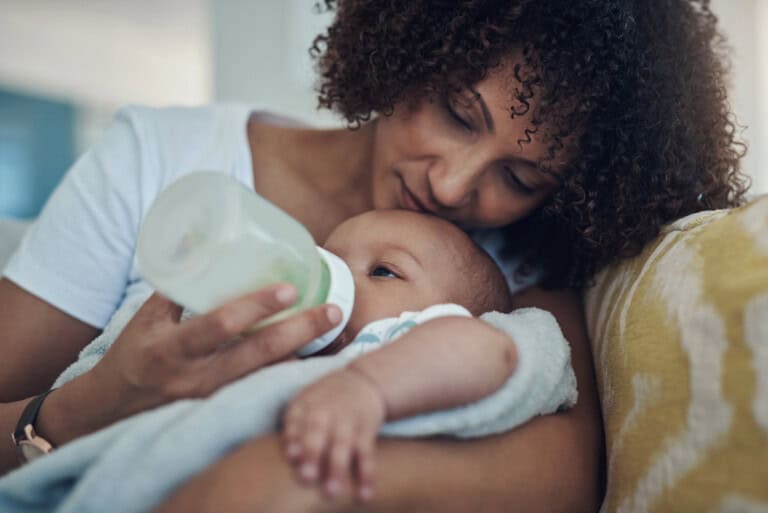 A mother with curly hair is experiencing silent struggles as she feeds her baby with a bottle while sitting on a couch. The infant, wrapped in a light-colored blanket, rests peacefully in the soft-lit indoor setting, unaware of the quiet challenges unfolding around them.