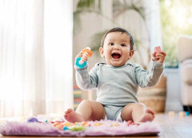 A baby wearing a light gray outfit is sitting on a soft, fuzzy rug, holding a toy in each hand. The baby is smiling widely. In the background, there are plants and natural sunlight coming through large windows. It's the perfect moment to ponder middle names while enjoying this serene scene.