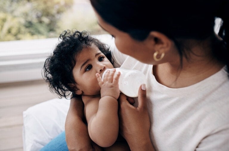 A person is bottle-feeding my baby, who has curly black hair and is gazing up at them. The person, adorned in a white shirt and gold hoop earrings, cradles the baby gently. The background is blurred, with a hint of greenery providing a serene backdrop to this tender moment.