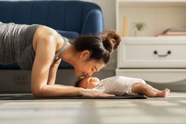 A woman engaging in postpartum fitness is doing a plank exercise while leaning down to touch noses with a baby lying on a yoga mat. The baby, dressed in a striped onesie, giggles as they bond. In the background, there's a blue couch and a white dresser.