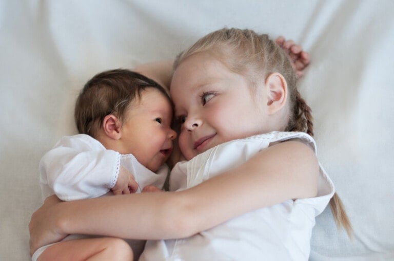 A young girl with braided hair lies on a white surface, embracing a newborn baby. Both children are wearing white clothing. The baby has dark hair and is looking at the older child, who is smiling gently, as if cherishing one of the sweet gifts from the new baby.