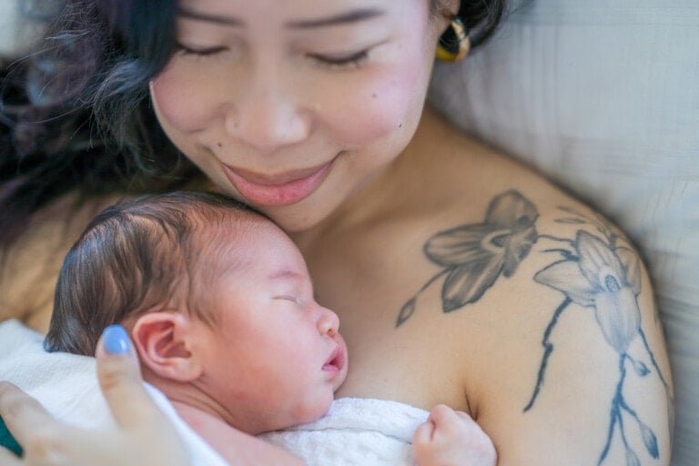 A woman with a floral tattoo on her shoulder is holding a sleeping newborn baby against her chest. She is smiling slightly with her eyes closed, exuding the serene contentment often associated with an orgasmic birth. The scene conveys a sense of warmth and bonding.
