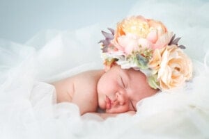 A sleeping baby, adorned with a large floral headband made of pink and peach flowers, rests on a white, lacy fabric background. It's a scene as serene and delicate as an elegant name whispered softly into the night.