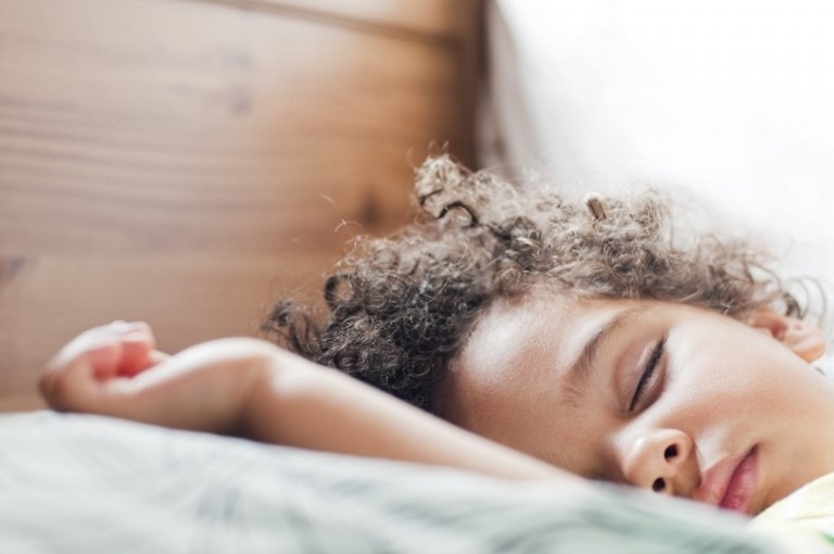 A child with curly hair is sleeping on their side, resting on a pillow. The background shows a wooden surface and some bright light coming from a window. Practicing good sleep hygiene can help achieve such peaceful and relaxed sleep for kids.