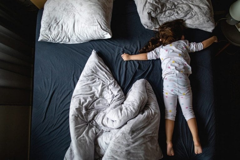 A child in pajamas is lying face down on a bed with outstretched arms, demonstrating unique sleep habits while traveling. The bed has a dark sheet and two pillows, with one pillow at the top and one on the side. A light-colored comforter is crumpled next to the child.