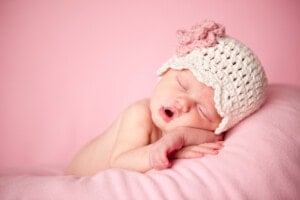A sleeping baby rests on a light pink blanket, wearing a white crocheted hat with a pink flower. The baby, who could be named Lily or Ava, is slightly positioned on their side with their mouth open. The background is a solid pink color.