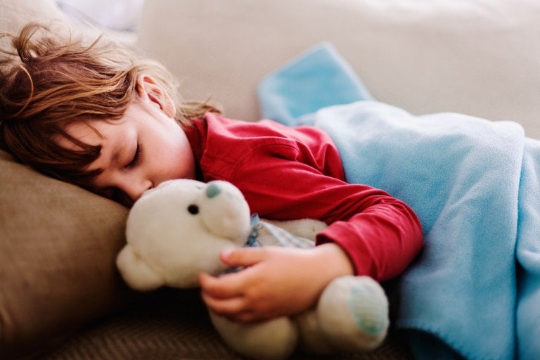 A young child with brown hair is sleeping on a beige couch, holding a white teddy bear. The child is wearing a red long-sleeve shirt and is partially covered by a blue blanket. The scene appears peaceful and cozy, perfect for prepping your baby for Spring Forward.