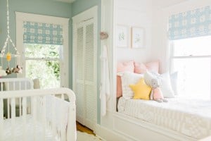 A bright, white-walled tiny nursery features a crib and a built-in bed nook. The crib has hanging toys, and the bed is adorned with pillows, a plush bunny, and a star pillow. The room is decorated with patterned window shades and has a closet with louvered doors.
