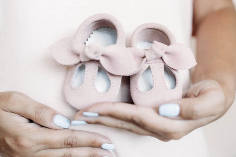 A person is holding a pair of small, pink baby shoes with bows in their hands. The background is light and blurred, emphasizing the focus on the shoes. With "it's a girl" written all over this moment, the person's light blue painted fingernails add a delicate touch.